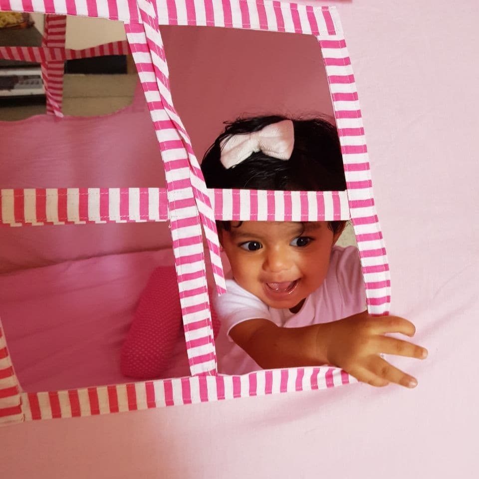 A baby laughing through the window of a pink striped play tent.